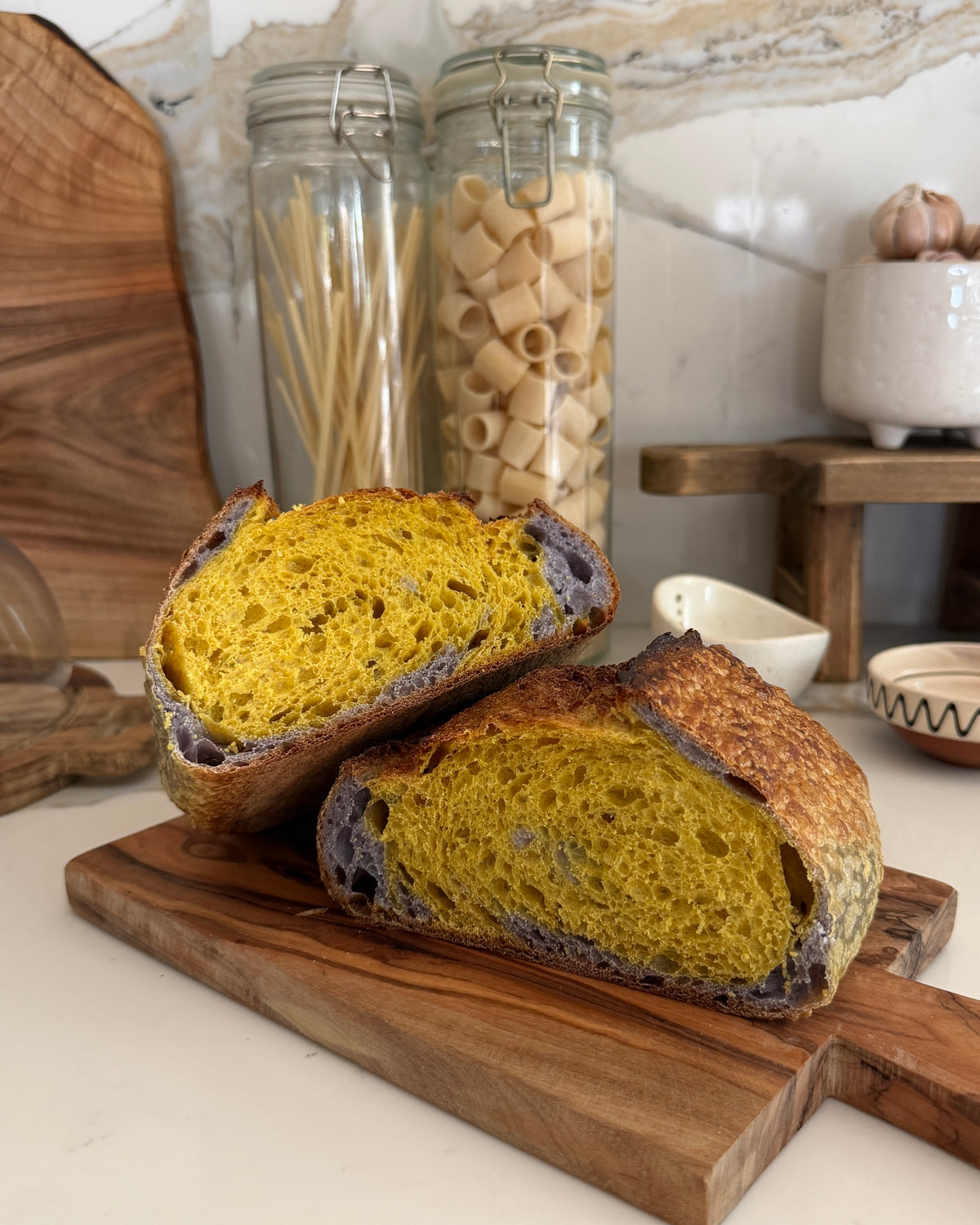 Two halves of Häsi sourdough bread loaf with a turmeric and a butterfly pea flower interior, displayed on a wooden cutting board in a kitchen setting with pasta jars in the background.