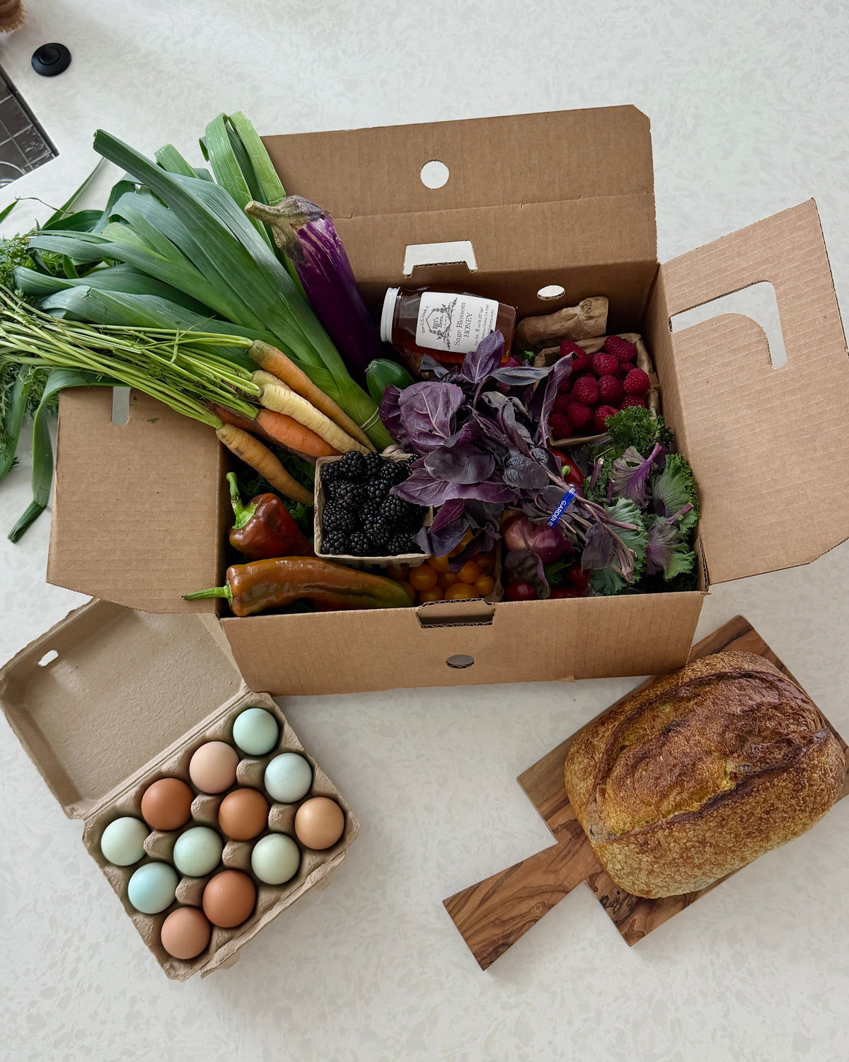The Full Market box from Market Mama filled with fresh produce, a jar of honey, and a small container of blackberries, arranged on a light countertop with a carton of eggs and a loaf of bread nearby.